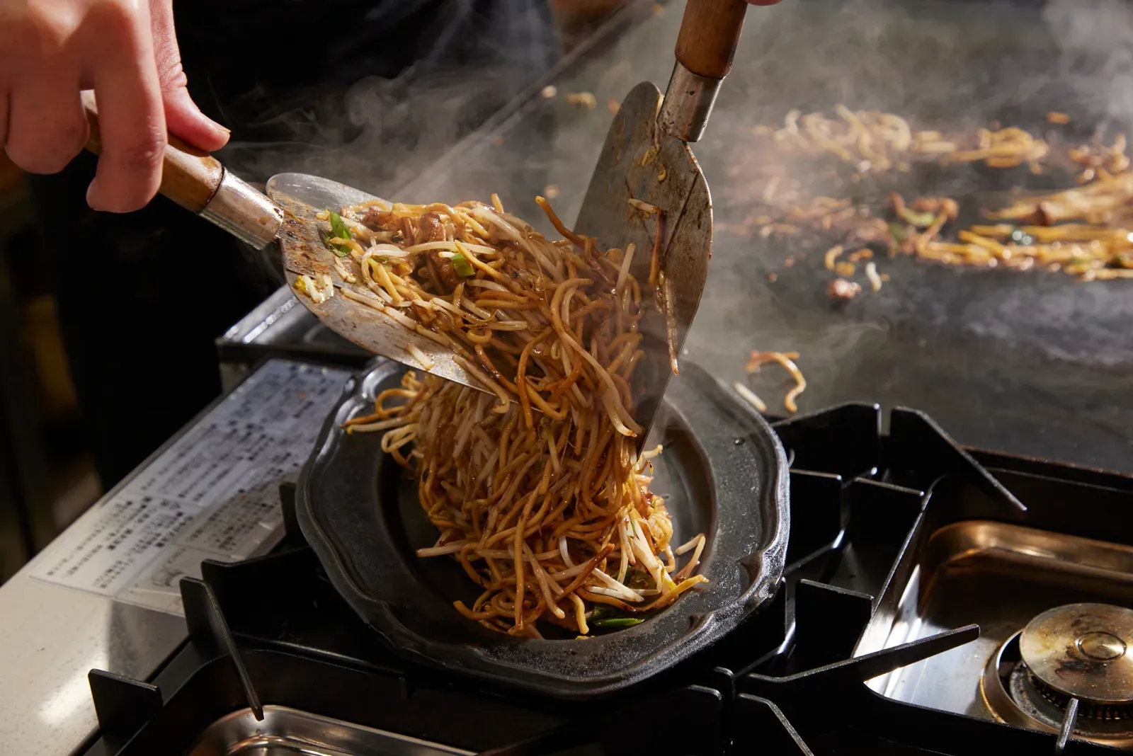 二枚のへらで麺を持ち上げ、鉄板で焼き上げる様子